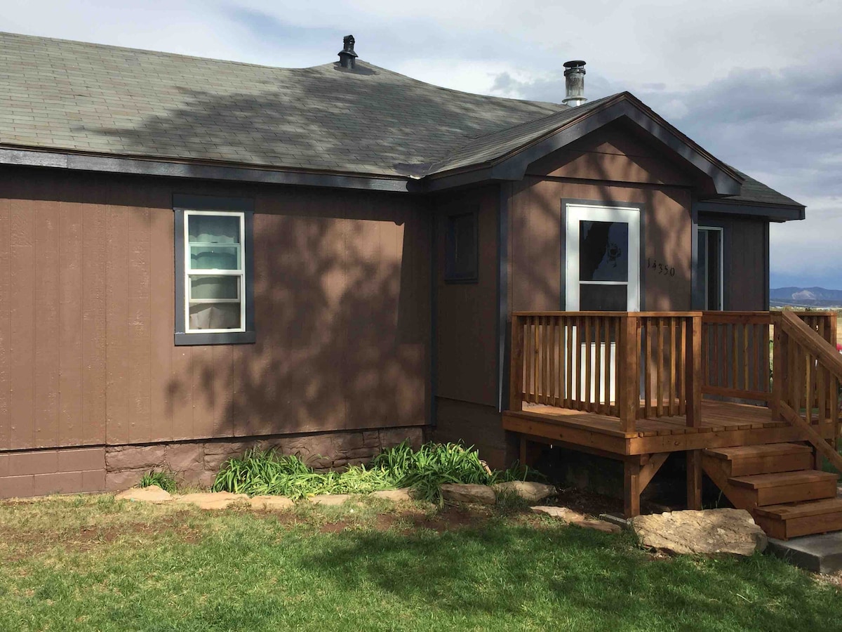 The image displays the exterior of a single-story home with a wooden deck and steps leading to the entrance. Surrounding the home, a green lawn and a few shrubs are visible. The structure features a brown wooden facade, with windows and a chimney atop the sloped roof.