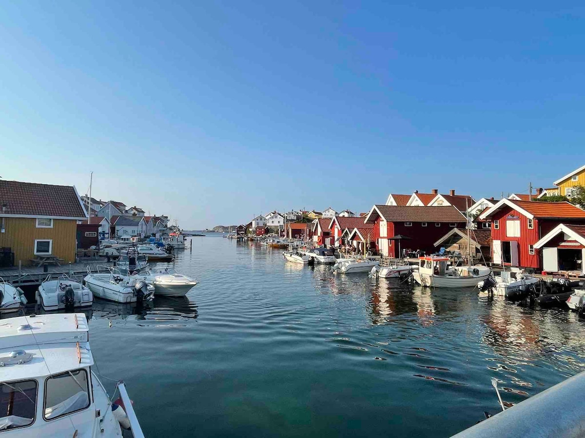 A calm harbor scene is depicted, featuring a narrow waterway lined with charming red houses. Several small boats are moored along the edges, reflecting the clear blue sky above. The peaceful atmosphere is enhanced by the stillness of the water.