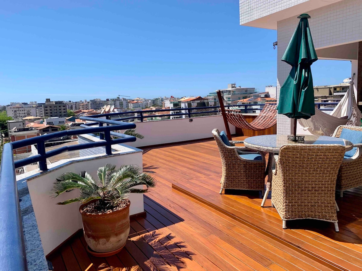 A spacious terrace features polished wooden flooring, complemented by a dining table with woven chairs. A large green umbrella provides shade, while a hammock is also present. The panoramic city view is visible in the background, highlighting a mix of residential buildings.