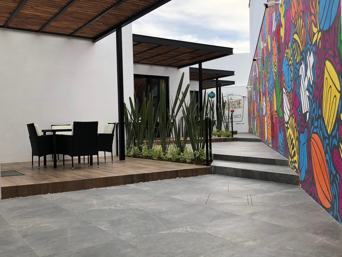 A shared outdoor area features a colorful mural on one wall and a wooden deck with a table and chairs. Lush greenery is planted alongside the path leading to the entrance, framed by modern structural elements and gently sloping steps.