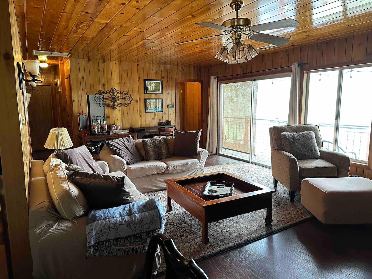 The living area offers a warm ambiance with wood-paneled walls and a ceiling fan. Seating includes a light-colored sofa and chairs, arranged around a coffee table on a large area rug. Natural light fills the space through glass sliding doors leading to the deck.