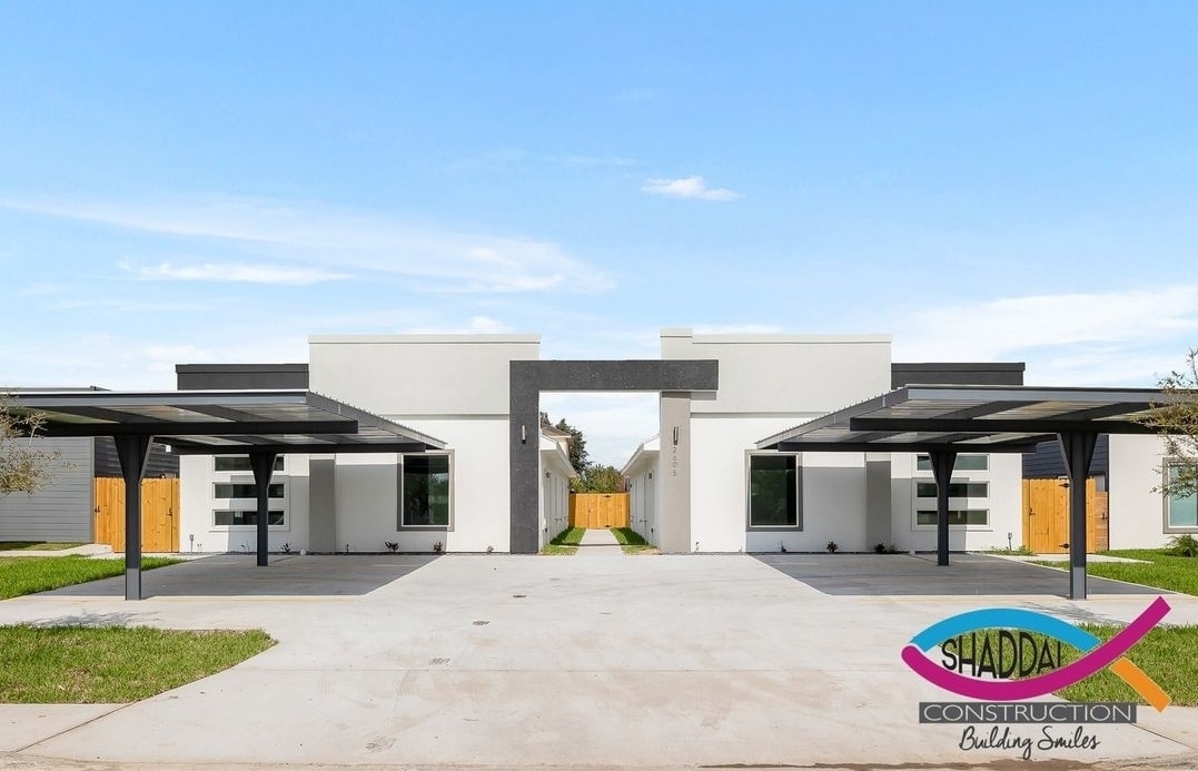 A modern building exterior is featured, showcasing two structures separated by a central pathway. Covered parking spaces are visible in front of each unit, with a neatly maintained lawn and a wooden fence in the background. Clear blue skies provide a bright and open atmosphere.