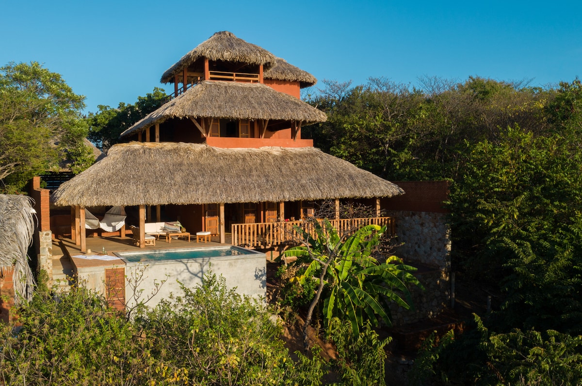 A multi-story house is enveloped by lush greenery, featuring a thatched roof and spacious terraces. A swimming pool is located on the ground level, with relaxed seating areas positioned around it, providing an inviting space for guests to unwind in nature.