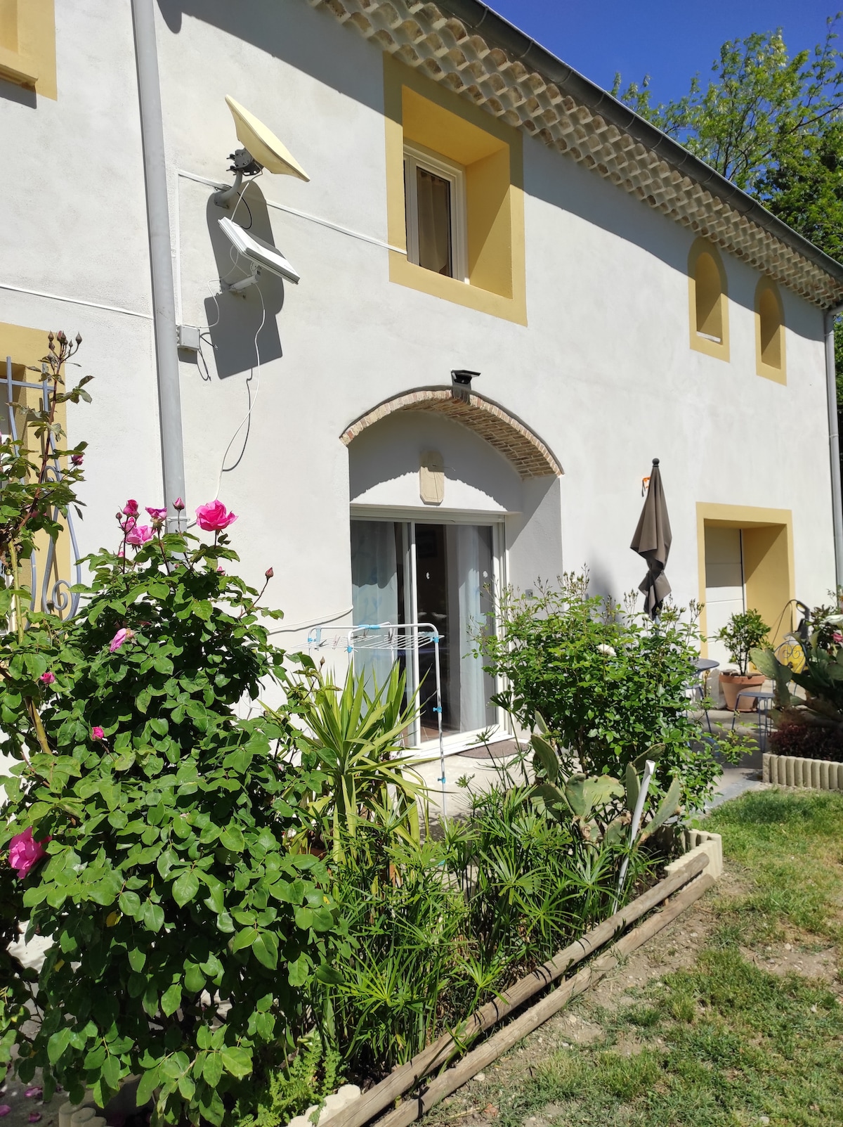 The exterior of the property features a white facade with yellow accents and several windows. A welcoming entrance is framed by greenery, including flowering plants. A patio area is visible, with a sunshade enhancing the outdoor space.