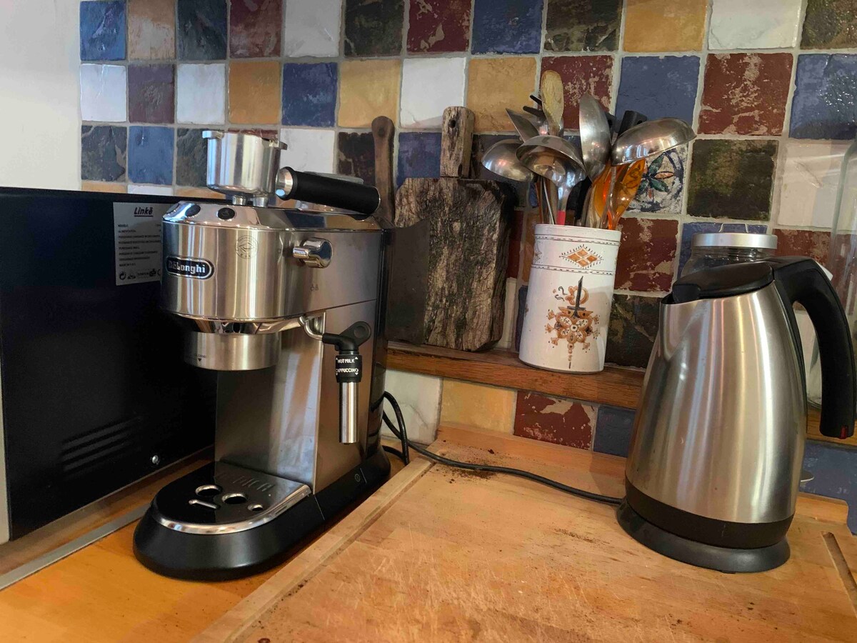 A modern coffee machine is positioned on a wooden countertop, accompanied by a kettle. Behind, an array of utensils is neatly organized in a container against a backdrop of colorful tiled walls. The space reflects a functional and inviting kitchen area.