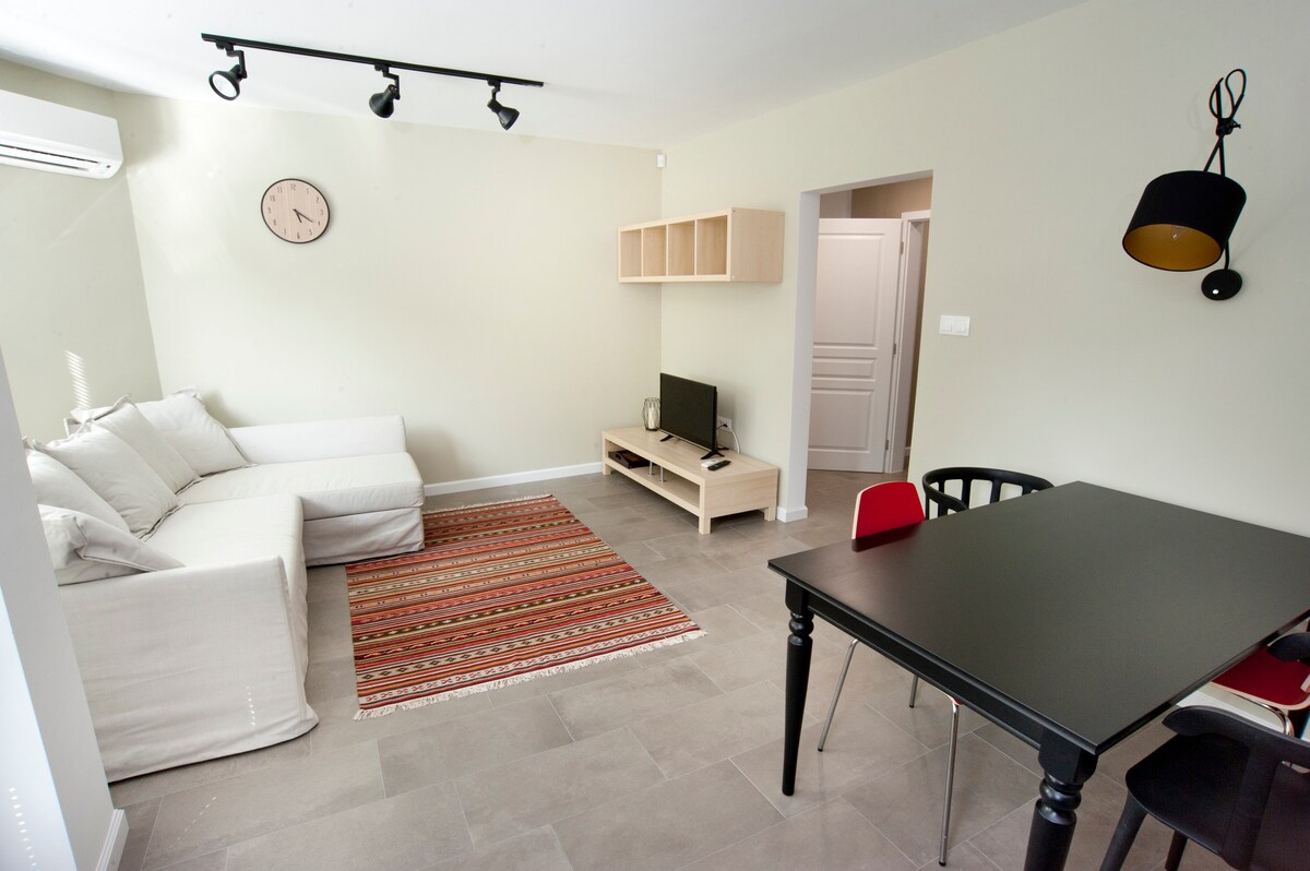 A spacious living area is equipped with a comfortable sectional sofa accompanied by a traditional rug. A wall-mounted clock and flat-screen TV are present, while a black dining table with red chairs offers a cozy dining space. Soft natural light illuminates the room.