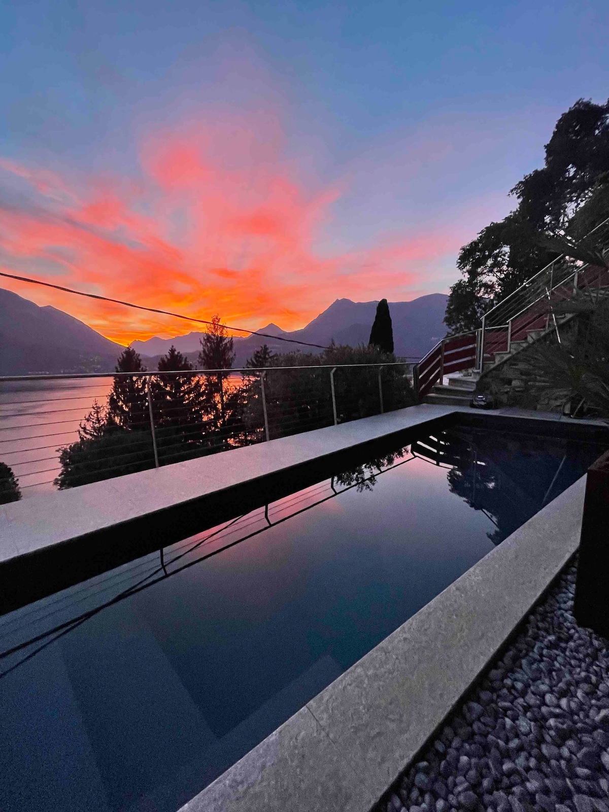 A tranquil swimming pool reflects the vibrant colors of a sunset over the mountains and lake. Lush greenery frames the scene, while a sleek railing enhances the contemporary design. The serene atmosphere invites relaxation amidst nature's beauty.