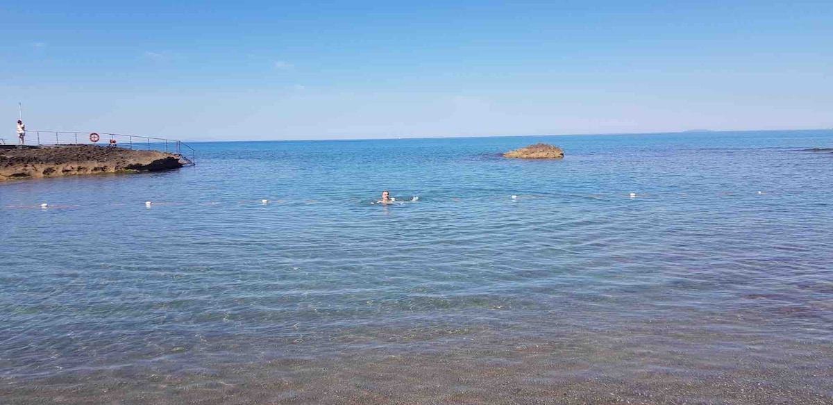 A serene seascape is presented, featuring calm waters with gentle ripples. A person is visible swimming in the distance, while a rocky formation emerges from the water. The clear blue sky complements the tranquil atmosphere and enhances the coastal view.