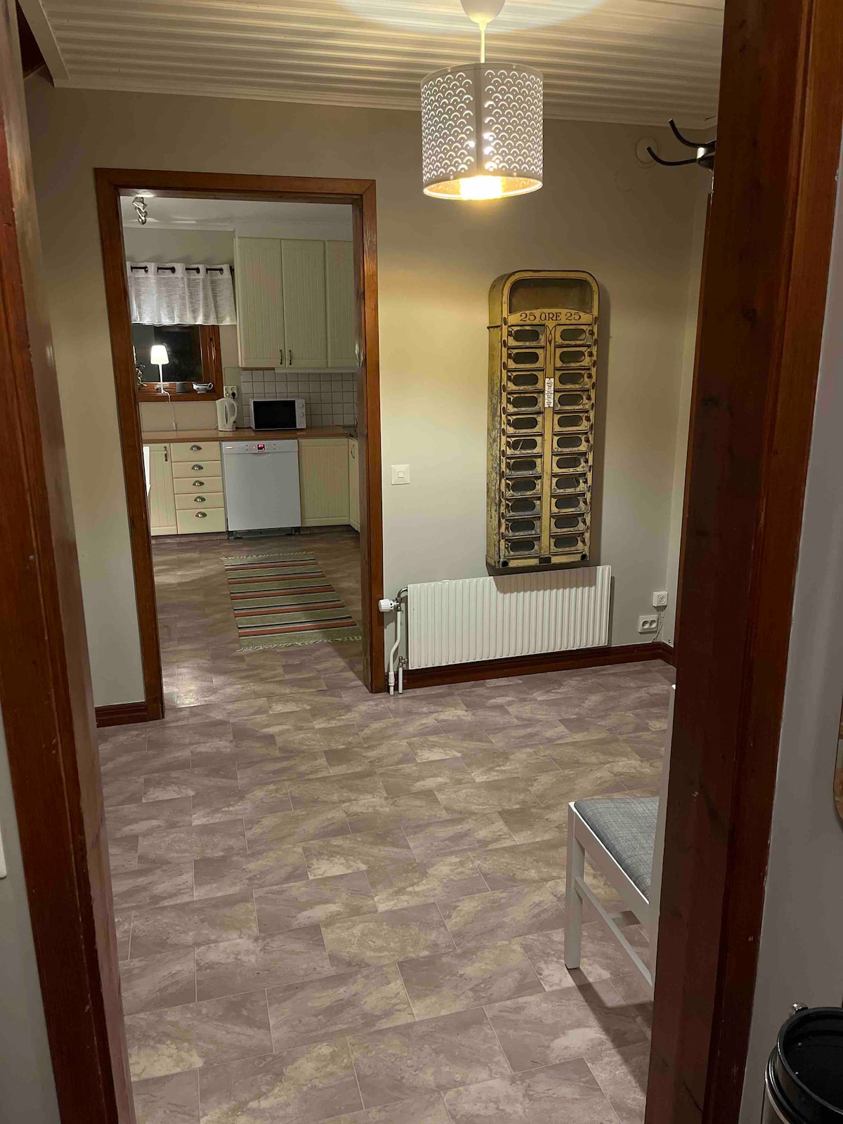 A hallway is presented, leading toward an open kitchen area. The walls are adorned with light-colored paint, and a pendant light hangs from the ceiling. A coat rack and a small chair are visible, while the flooring features a subtle patterned design.