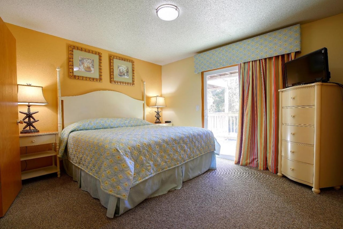 A brightly colored bedroom features a spacious bed dressed in a cheerful yellow-patterned coverlet. Complementary bedside lamps are positioned on either side, and a tall dresser stands nearby. Natural light enters through sliding glass doors that lead to a small outdoor area.