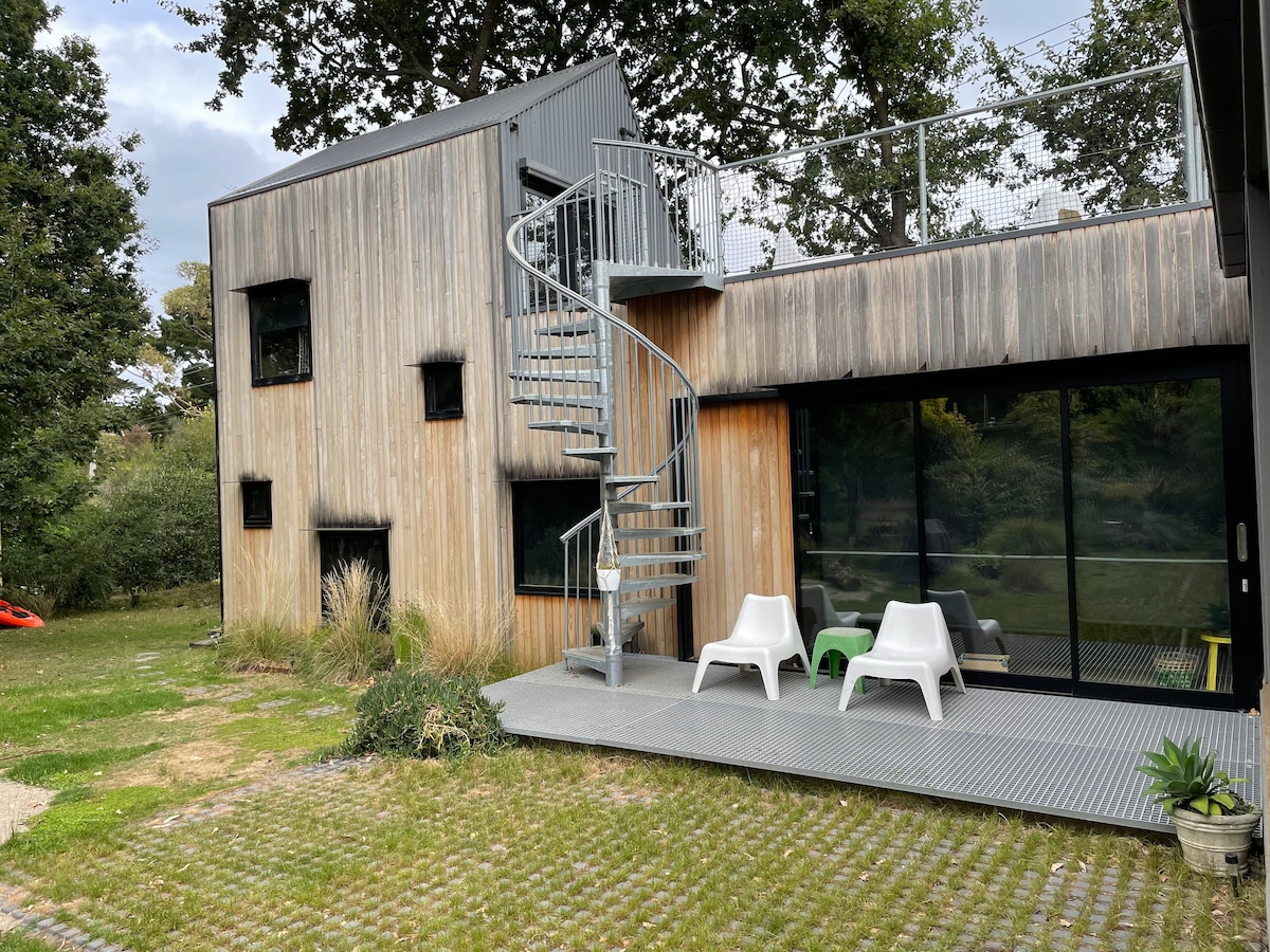 A modern two-story building is shown, featuring wooden siding and large glass windows. A spiral staircase leads to an upper level, where outdoor seating is visible on a metal deck. The surrounding area is landscaped with grassy patches and plantings.