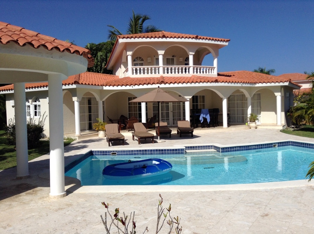 A spacious villa with a red tile roof is surrounded by tropical gardens. A private swimming pool is visible, featuring a circular lounge area. Several sun loungers are arranged by the poolside under a large umbrella, and large glass doors lead to an inviting terrace.