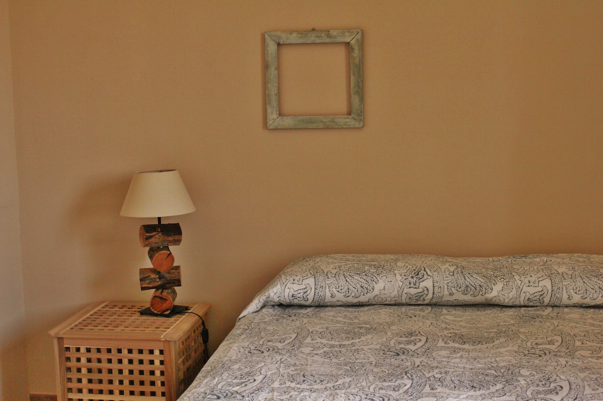 A spacious bed covered with a patterned duvet is positioned against a neutral wall. Beside the bed, a wooden lamp with a light shade rests on a decorative nightstand. A simple empty frame hangs above the headboard, adding an understated decorative touch.