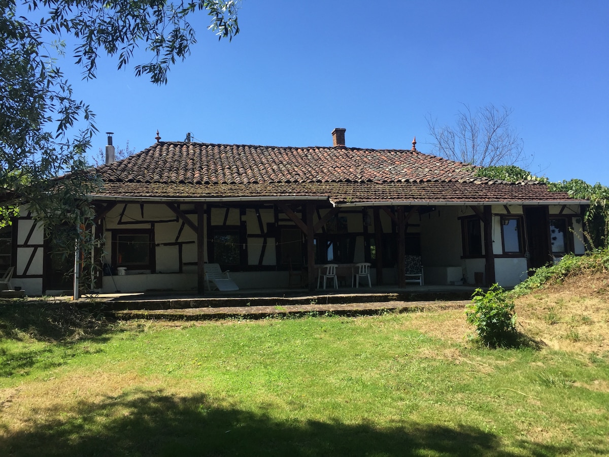 A traditional half-timbered house is set against a clear blue sky, surrounded by grassy areas. The front porch features a few chairs, providing a space for relaxation. Lush greenery borders the property, enhancing the natural ambiance of the tranquil setting.