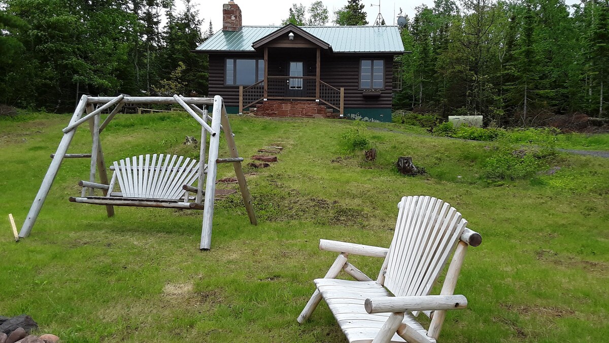 A charming cabin is visible in the background, framed by lush greenery. A wooden swing set is positioned beside two Adirondack chairs on a well-maintained lawn. The front steps lead to a welcoming porch, creating an inviting atmosphere for relaxation outdoors.