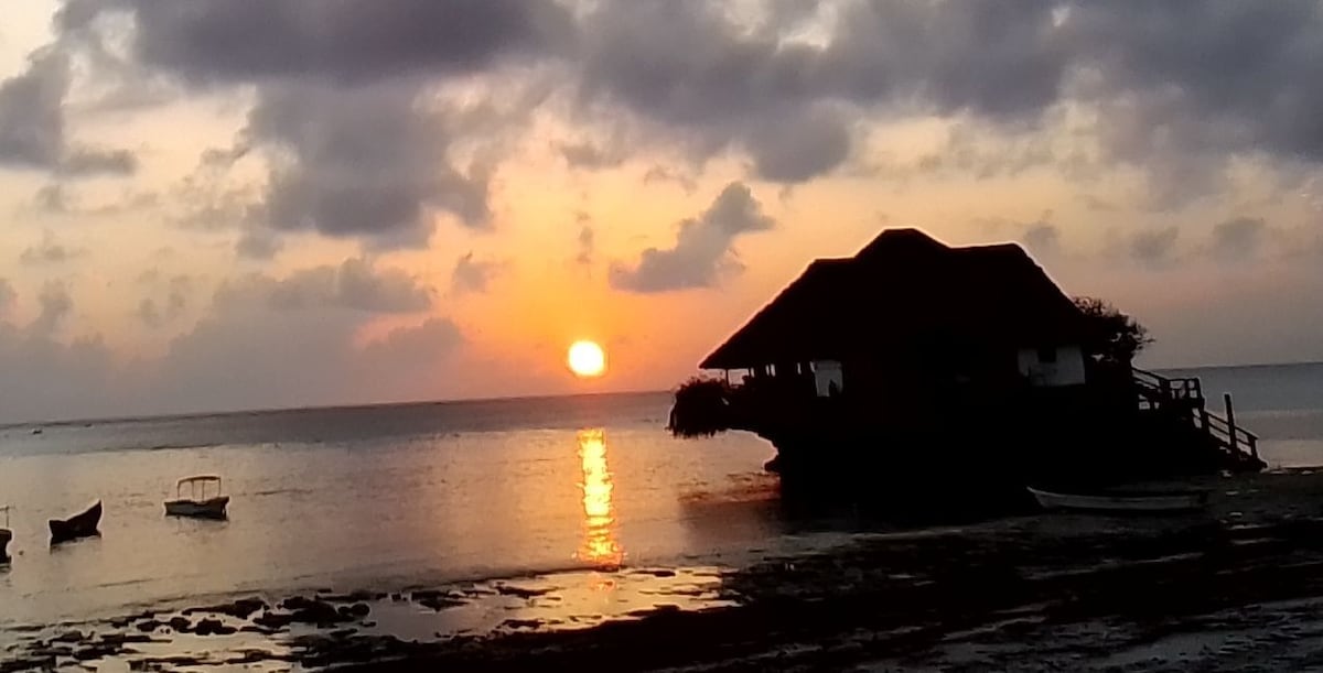A silhouette of a thatched-roof house is positioned against a vibrant sunset over the ocean. The sun reflects on the water, creating a shimmering path. Small boats are visible in the foreground, adding to the serene coastal scene.