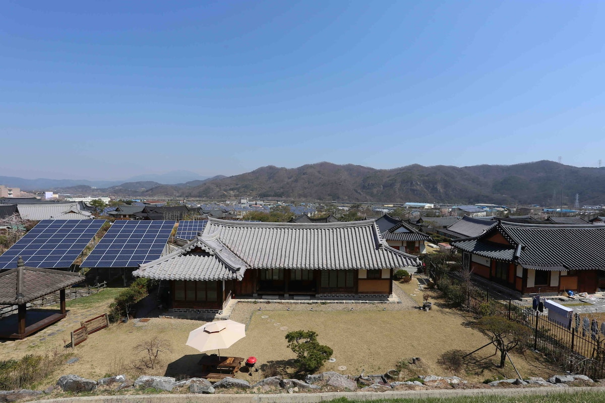 A traditional hanok is set against a clear blue sky, surrounded by rolling hills. Solar panels are visible nearby, while a grassy yard features a small umbrella and various plants. The architectural style emphasizes natural materials, blending harmoniously with the landscape.