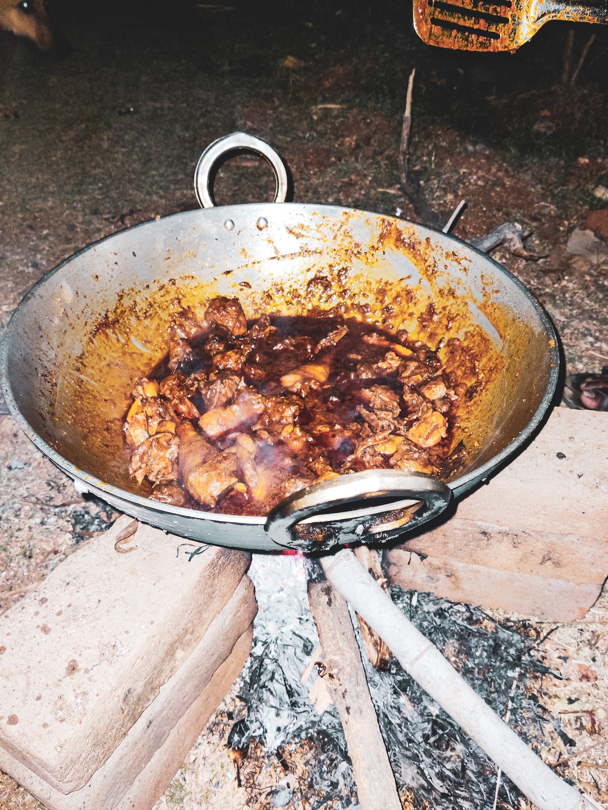 A large, round cooking vessel sits atop a makeshift stand, filled with a dark, savory dish. The pot shows evidence of a rich, simmering meal, with ingredients and spices evenly blended. The surrounding area features a rustic, outdoor setting with visible embers.