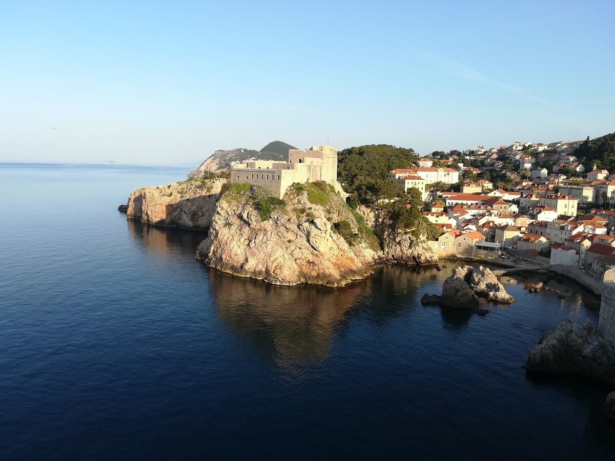 A coastal view captures a historic building perched on a cliff surrounded by calm waters. The skyline features a series of rooftops in warm tones, nestled against a backdrop of rolling hills and blue sky, offering a serene ambiance of the Adriatic coastline.