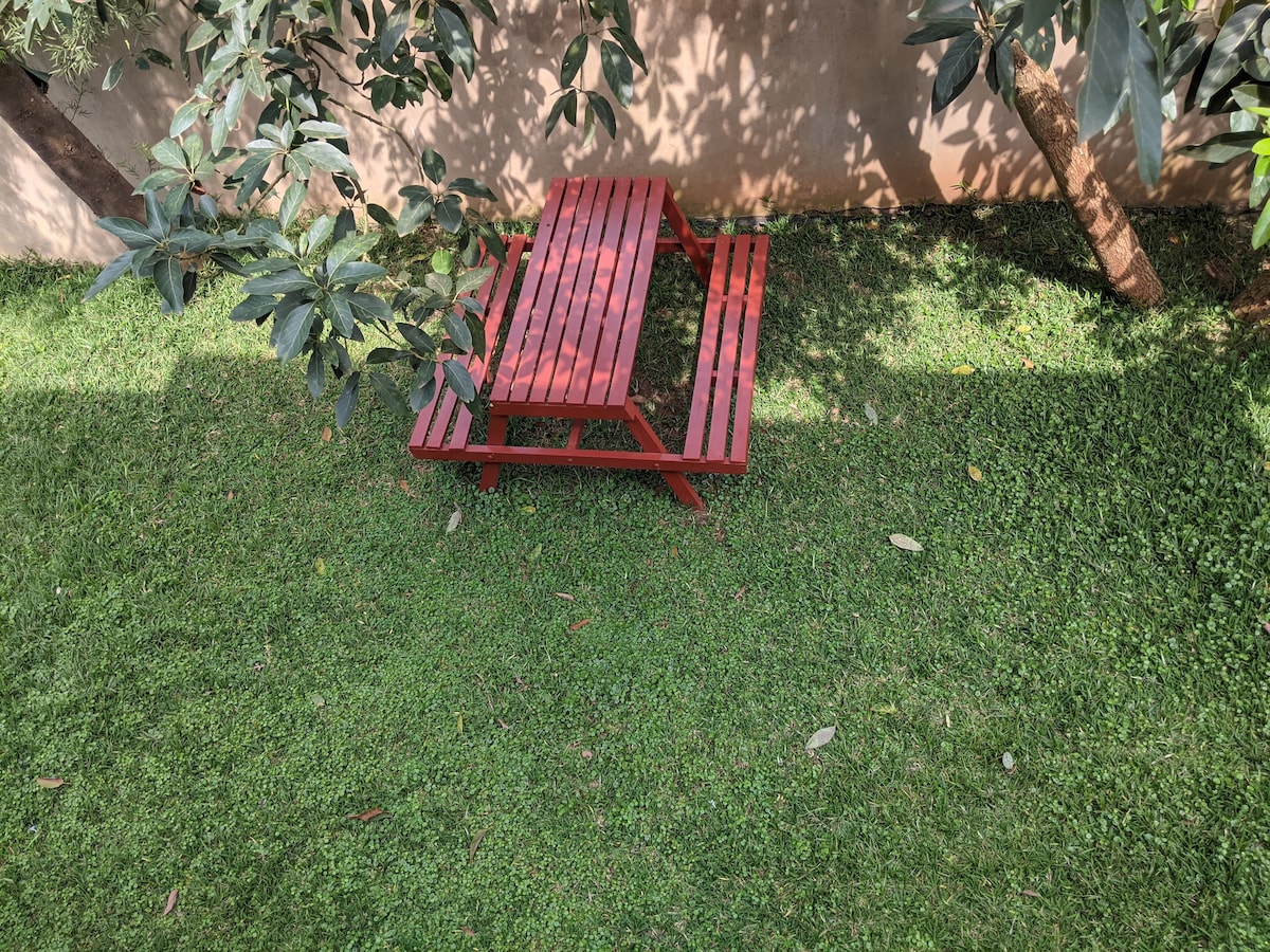 A red picnic table is positioned on a lush green lawn, surrounded by foliage. The smooth lines of the table create a contrast against the vibrant grass, while dappled sunlight filters through nearby leaves, casting soft shadows on the ground.