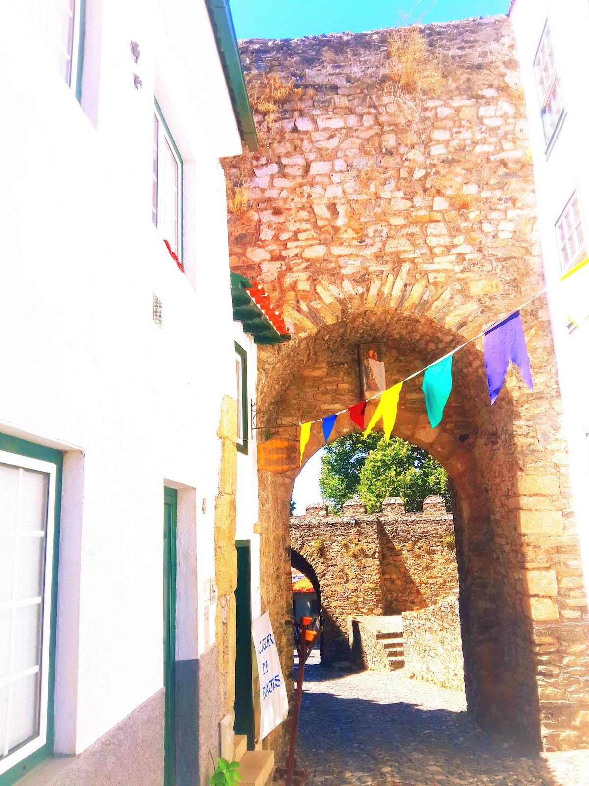 An ancient stone archway connects two white-walled buildings adorned with colorful flags. A narrow path leads through the arch, revealing a glimpse of a historic stone wall and greenery in the background, highlighting the rich history of the area.