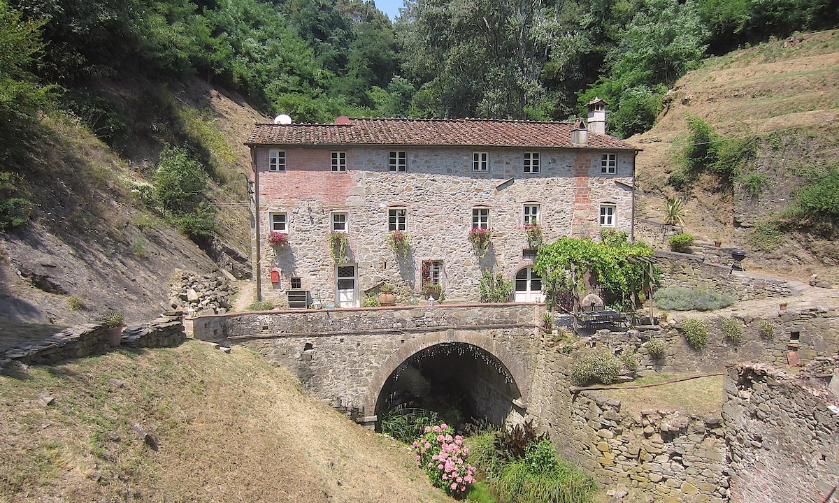 A historic stone house is set against a backdrop of lush greenery, with a charming bridge crossing over a small stream. The structure features multiple windows adorned with flower boxes. The surrounding landscape appears warm and inviting, emphasizing the serene and natural environment.