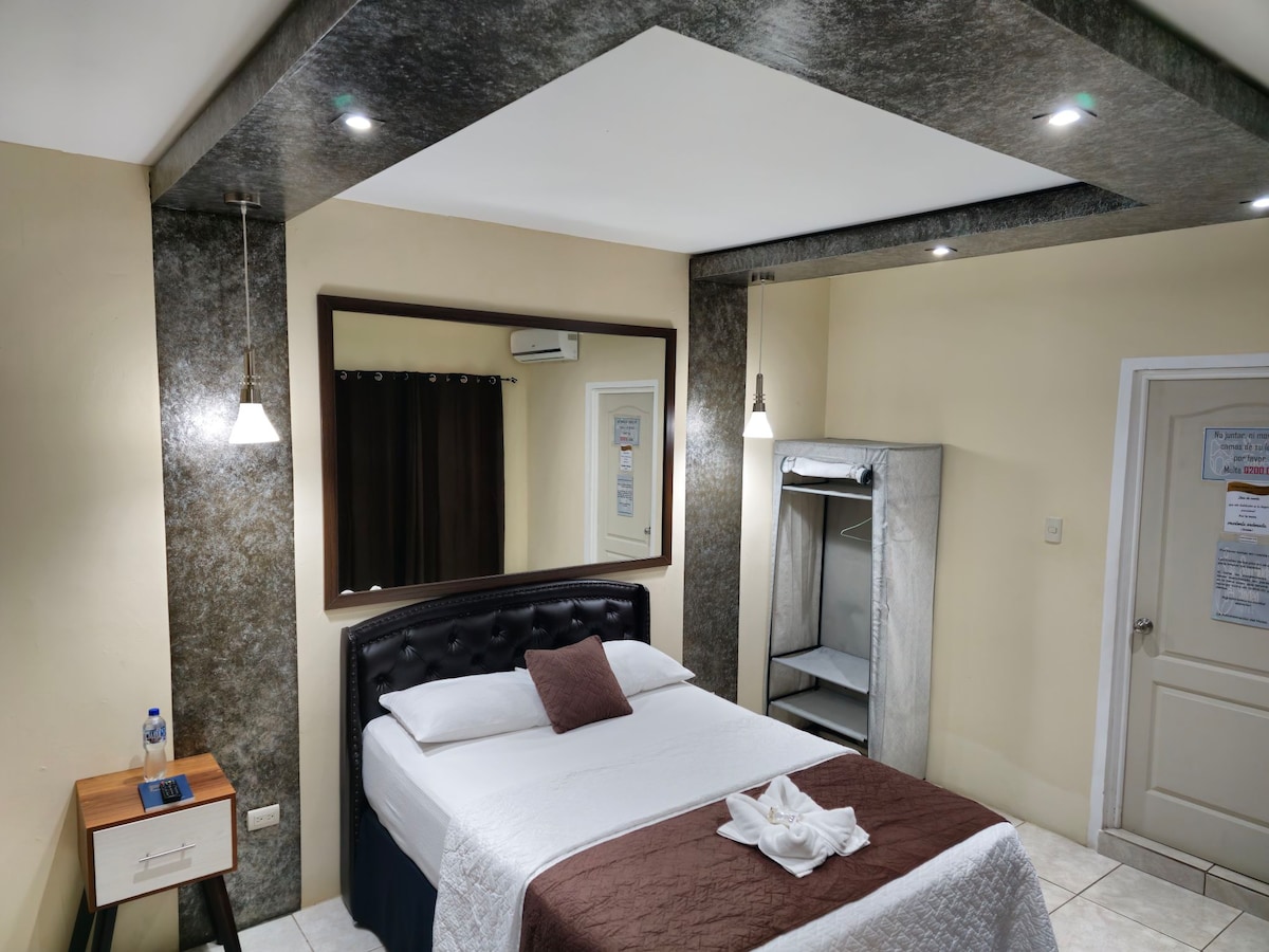 A neatly arranged guest room is presented with a spacious bed adorned with a light blanket and a decorative towel. The room features pendant lights, a large mirror, and a wardrobe, while neutral tones create a calming environment.