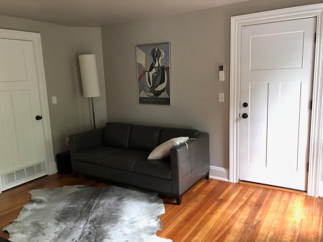 A modern living area features a sleek gray sofa facing a wall adorned with abstract artwork. The hardwood floor is complemented by a textured area rug. A floor lamp stands nearby, and a door provides access to the exterior.