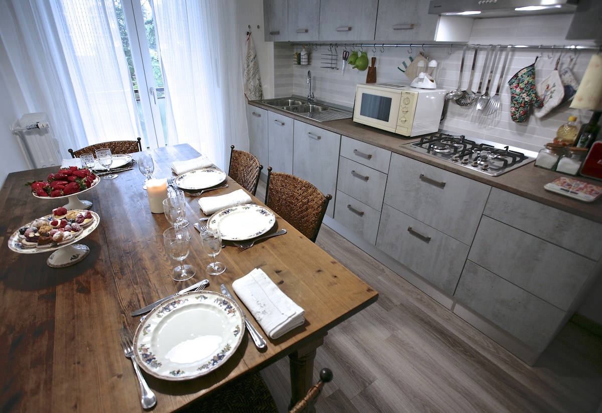 A functional kitchen area is depicted, featuring a wooden dining table set with plates and glasses. The cabinetry displays a neutral tone, complemented by essential appliances, including a microwave and a gas stove. Natural light filters in through sheer curtains, enhancing the space.