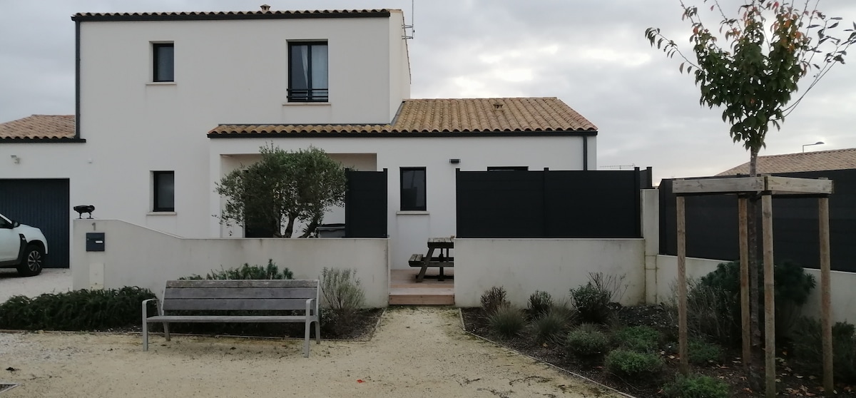 A white two-story structure with a tiled roof is visible, surrounded by a landscaped area. A wooden bench and a picnic table are situated in the outdoor space, alongside shrubs and greenery. A dark fence encloses the entrance, providing a private ambiance.