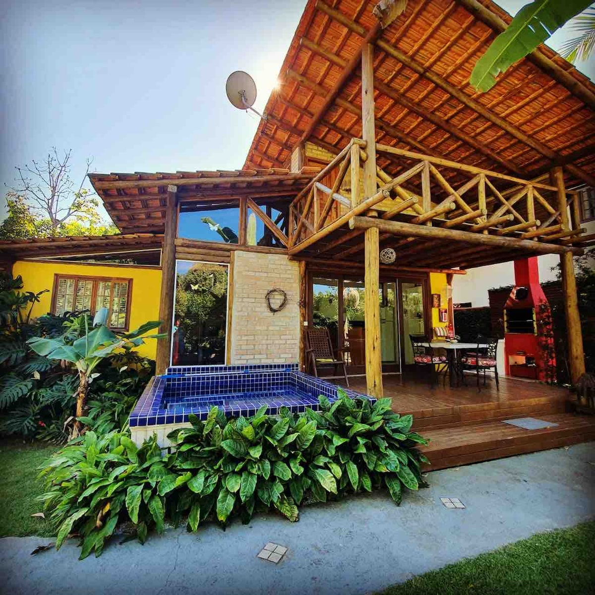 A two-story bungalow is visible, featuring a thatched roof and large glass doors that open to a wooden deck. Surrounding lush greenery and tropical plants enhance the outdoor space, while a small plunge pool adds a refreshing element to the patio area.