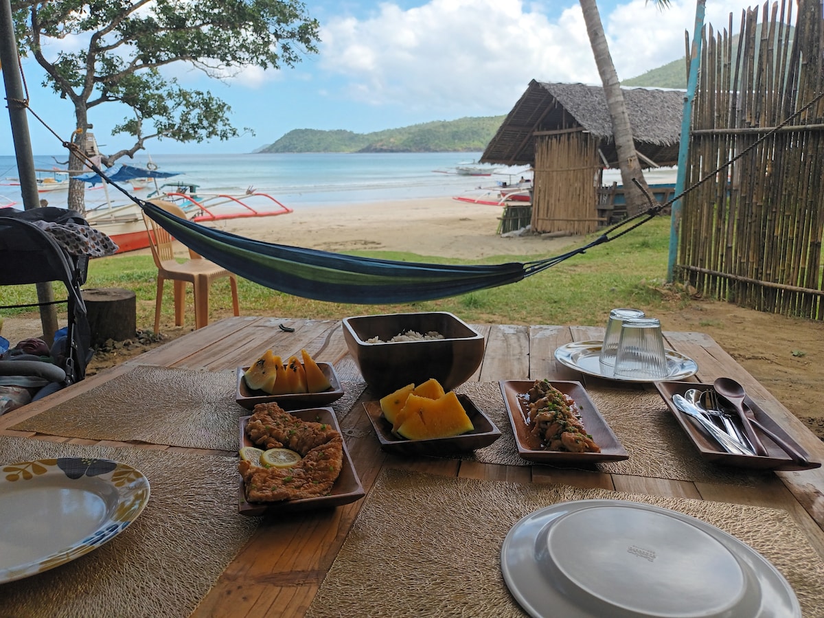 A wooden table showcases an assortment of food, including fresh fruits and grilled dishes, with dishes arranged neatly on brown trays. In the background, a view of the beach and ocean is visible, framed by palm trees and a rustic hut, under a partly cloudy sky.