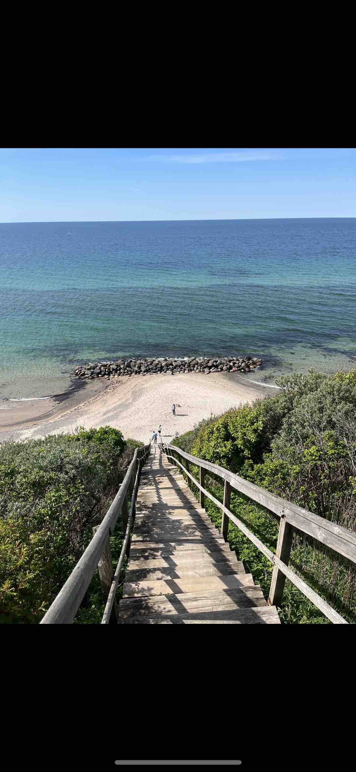 A wooden staircase leads down to a sandy beach, flanked by lush greenery. Calm blue waters extend towards the horizon, with a rocky outcrop visible offshore. A person is seen walking along the shore, illustrating the beach's accessibility and natural beauty.
