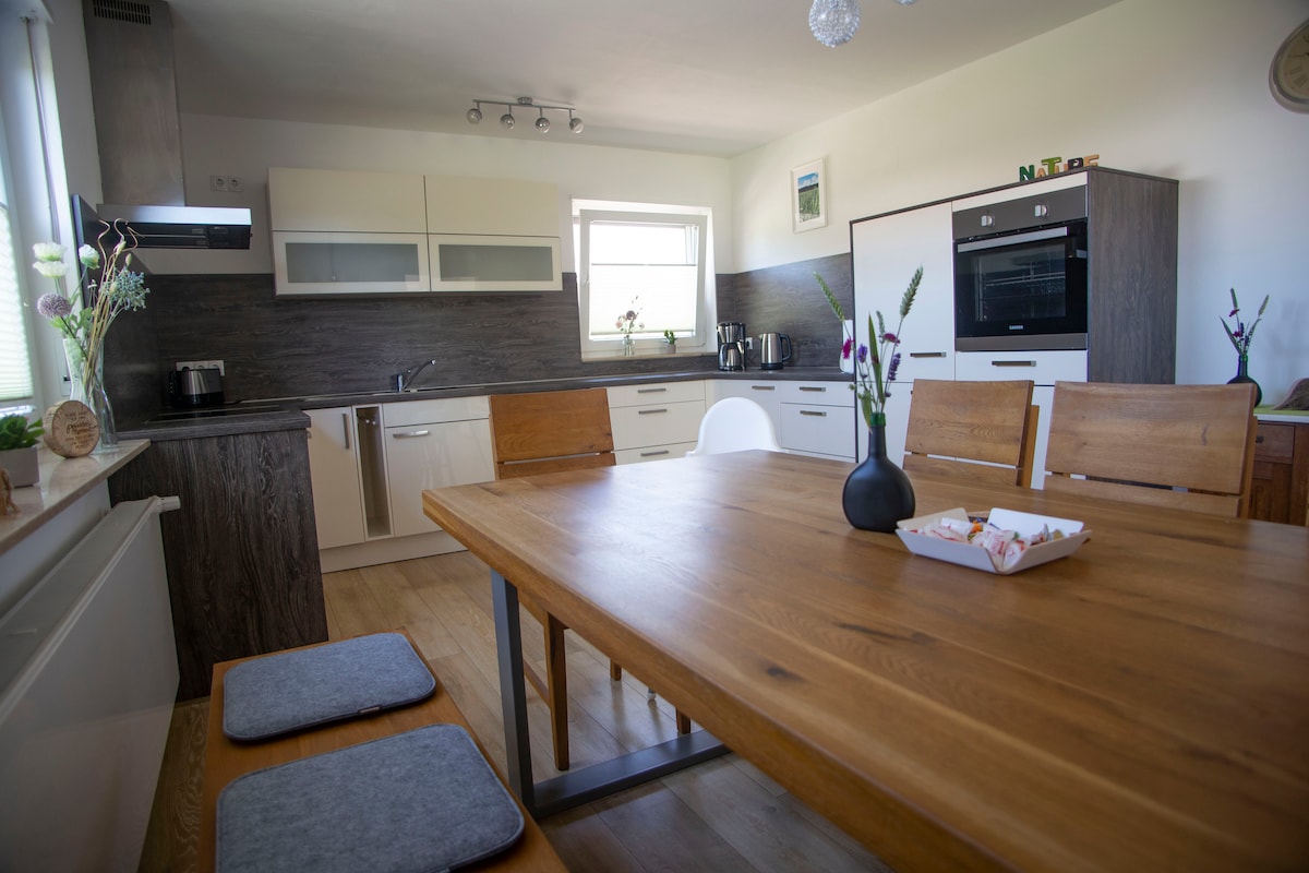 A modern kitchen is fitted with sleek cabinets and appliances. A wooden dining table is set with simple tableware. Two padded stools complement the table, while light streams through a window, illuminating the space.