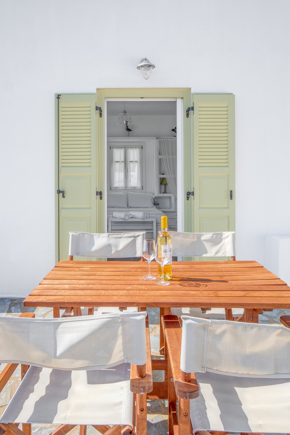 An outdoor dining area is presented with a wooden table and four folding chairs. Two glasses and a bottle of wine rest on the table. A door with green shutters leads into a bright interior space, enhancing the connection between indoor and outdoor living.