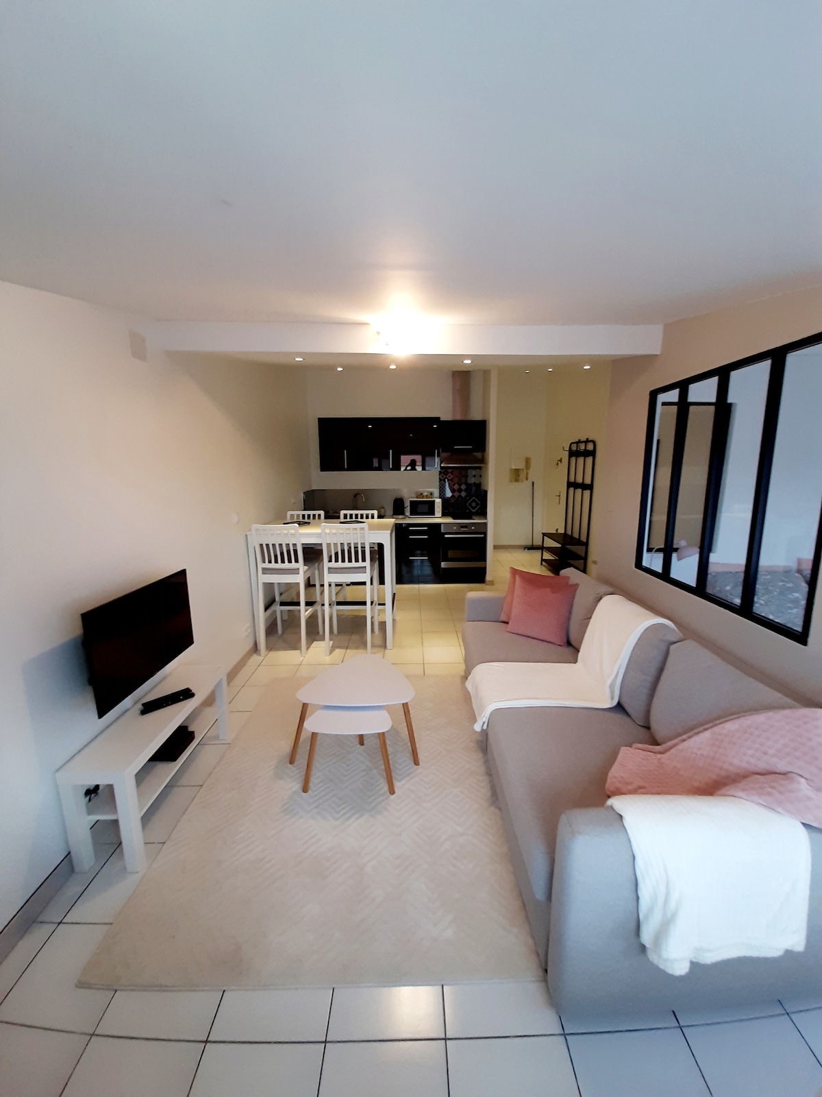The living area is designed with a light color palette, featuring a cozy sofa and a small coffee table. A dining space with four chairs is positioned next to a modern kitchen. Natural light filters through large windows, enhancing the open and airy atmosphere.
