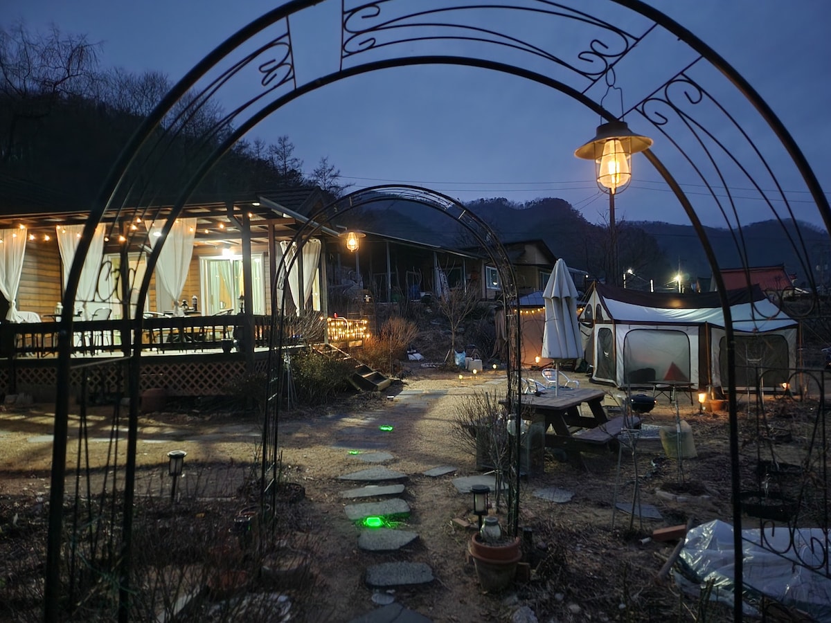 An outdoor space features an archway with a hanging lantern. Pathway stones lead towards a wooden structure with illuminated curtains. A communal seating area, surrounded by soft lighting, is visible along with a tent setup, creating a welcoming environment for gatherings.