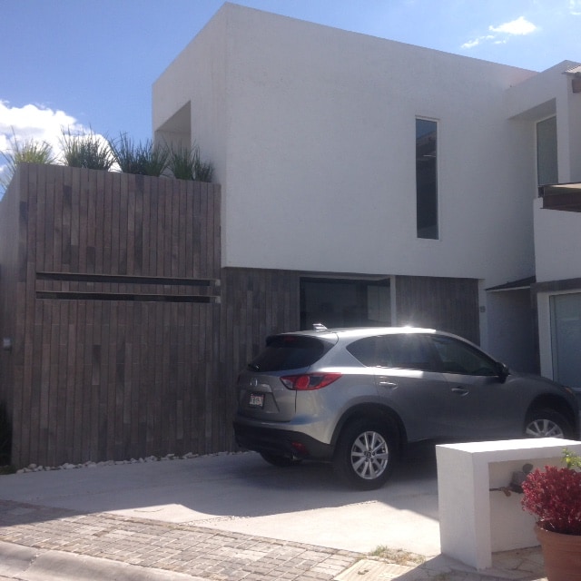 A modern two-story building is displayed, featuring a combination of smooth white and wooden-textured surfaces. A silver vehicle is parked on the driveway. Lush plants are visible atop a wooden fence that provides privacy while enhancing the exterior landscape.