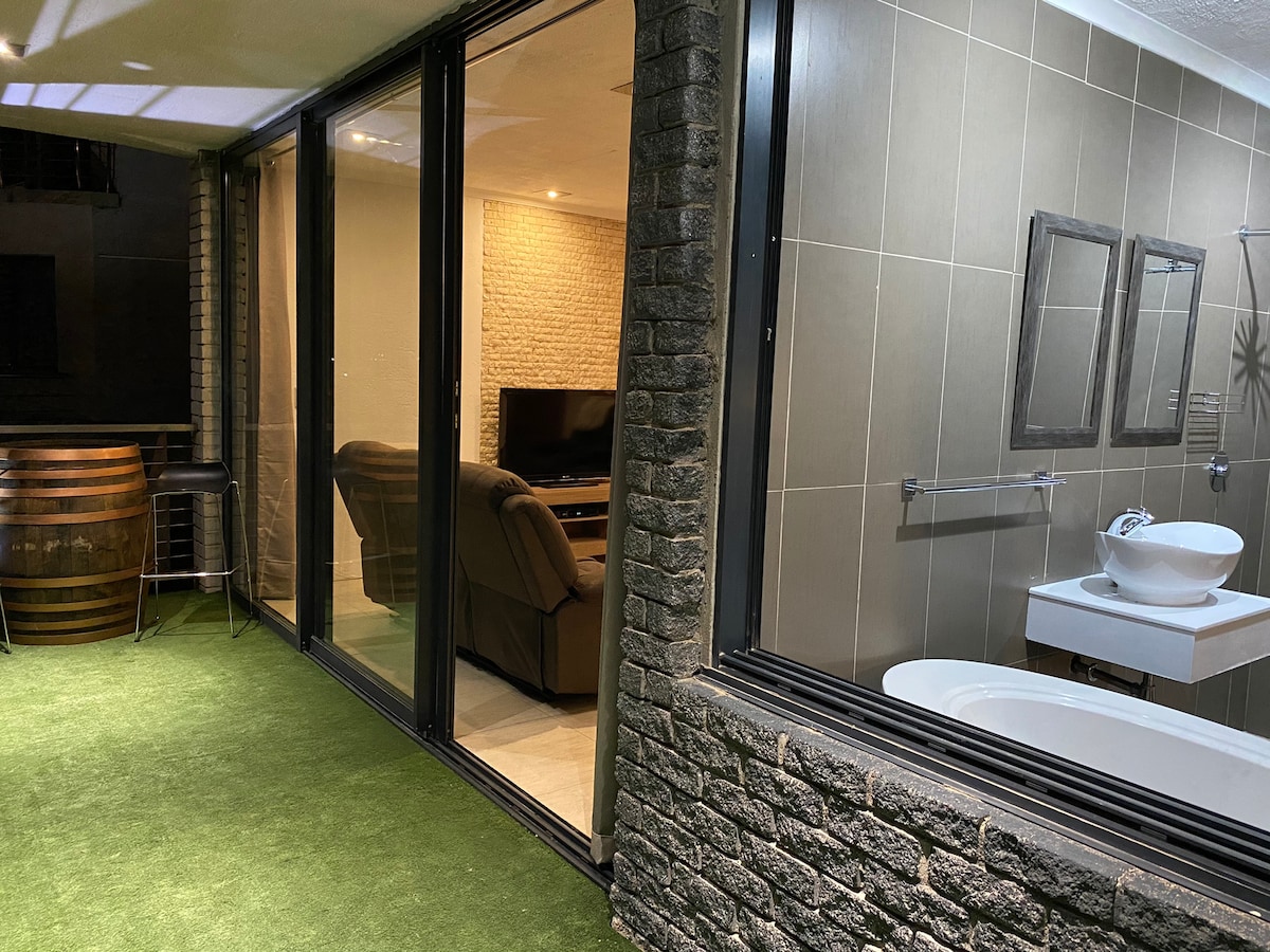 A private outdoor patio is visible, bordered by a stone wall. Large glass doors offer a view into a cozy lounge area with a flat-screen TV and comfortable seating. A modern bathroom design is highlighted, featuring a bathtub and a sink with mirrors.