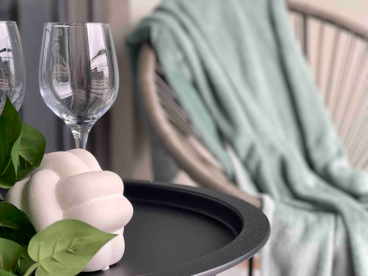 A balcony corner features a round black table adorned with a decorative object and two clear glasses. A soft, mint-colored blanket is draped over a comfortable chair, complemented by a green plant nearby, creating a serene outdoor space.