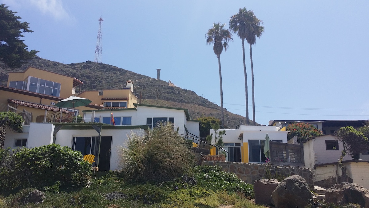 A coastal property is seen with multi-level structures showcasing colorful accents. Lush greenery surrounds the area, with large palm trees swaying gently. A mountainous backdrop and antennas are visible against a clear blue sky, creating a tranquil seaside atmosphere.