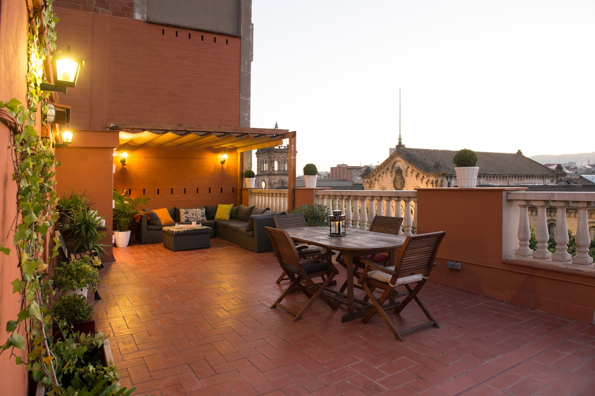 A spacious terrace features a comfortable seating area with a sofa, adorned with potted plants and decorative lighting. A wooden dining table with matching chairs is positioned for outdoor meals, providing panoramic views of the cityscape, including landmark buildings and distant hills.