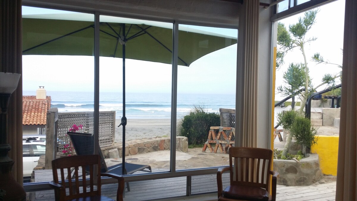 A large window offers a clear view of the sandy beach and ocean waves just beyond, framed by light-colored curtains. A green patio umbrella and outdoor seating are visible on the deck, inviting relaxation and enjoyment of the beach scenery.