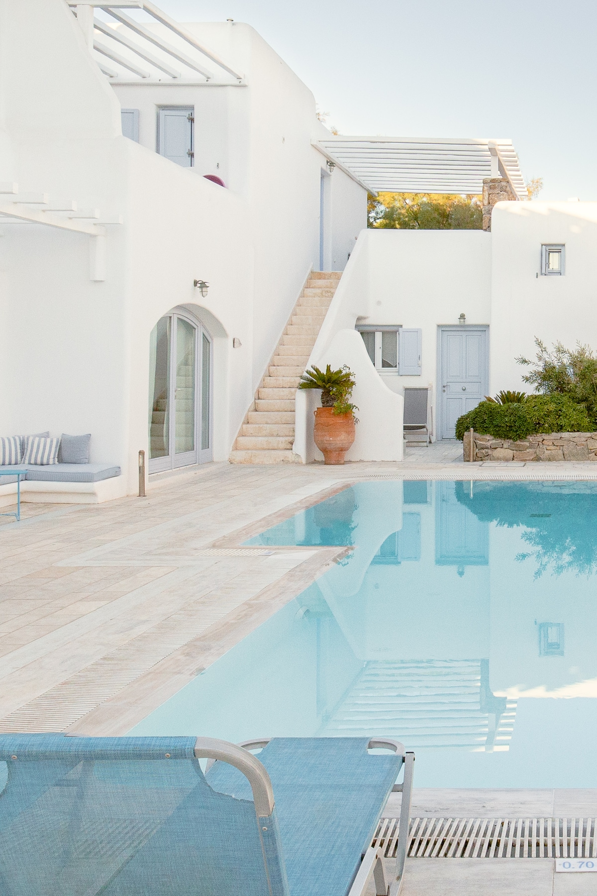 A serene view of a shared swimming pool surrounded by a light-colored stone deck, leading to an entrance of a villa. Whitewashed walls and blue accents are visible, along with a staircase. A lounge chair is positioned beside the pool, inviting relaxation.