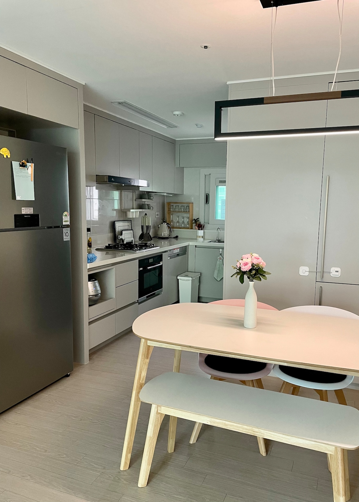 A modern kitchen is equipped with a refrigerator, oven, and stove, surrounded by sleek cabinetry. A dining table with three chairs sits in the foreground, featuring a small vase with flowers. Natural light illuminates the space through a window.