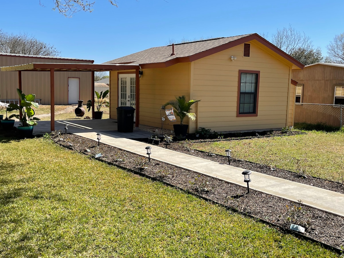 A small, yellow house is set against a clear blue sky, featuring a welcoming pathway lined with solar lights and bordered by mulch and plants. A covered porch is visible to the left, with a shadowed area providing additional outdoor space.