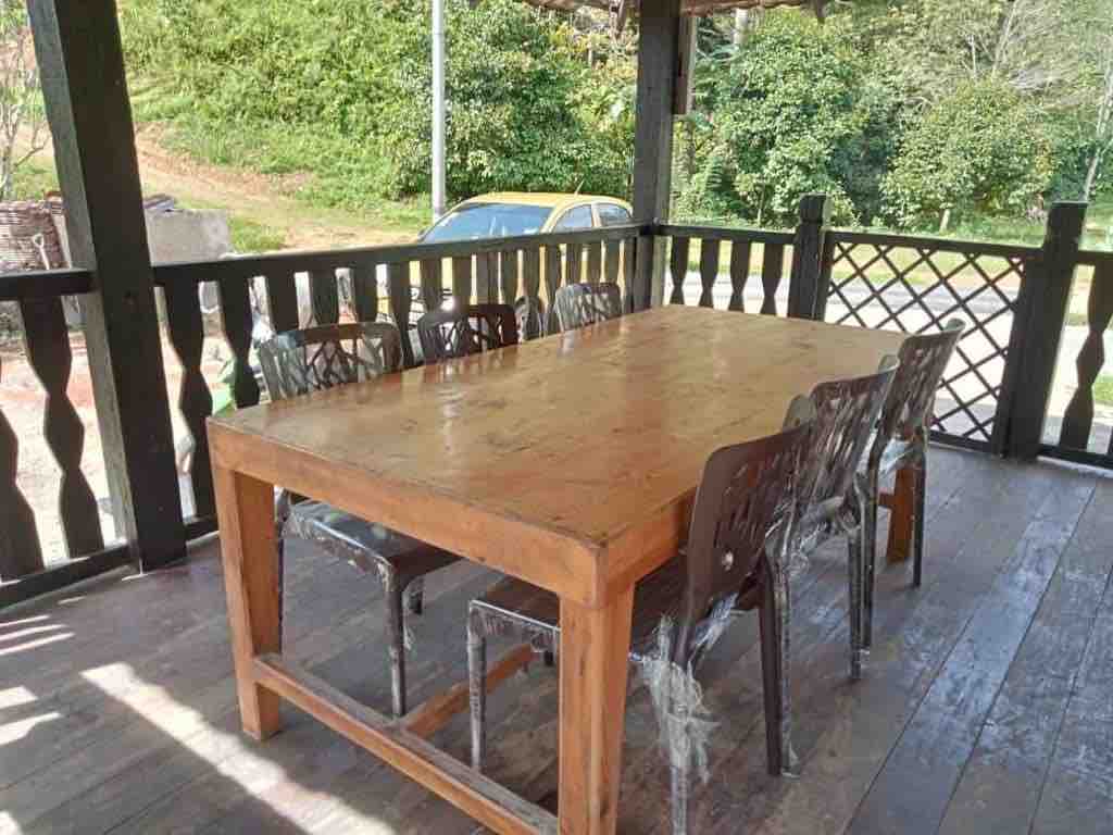 A large wooden dining table sits under a covered terrace, surrounded by six transparent chairs. The setting offers views of a verdant landscape, with greenery visible in the background. Sunlight casts a warm glow on the polished surface of the table.