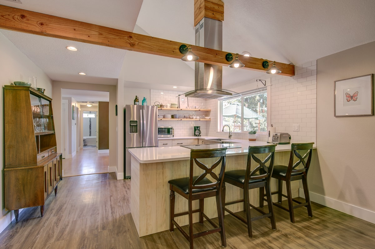 The kitchen features an island with seating for three, complemented by a modern range hood above. Stainless steel appliances are arranged along the sleek countertop, with ample natural light coming through large windows that provide views of the backyard.