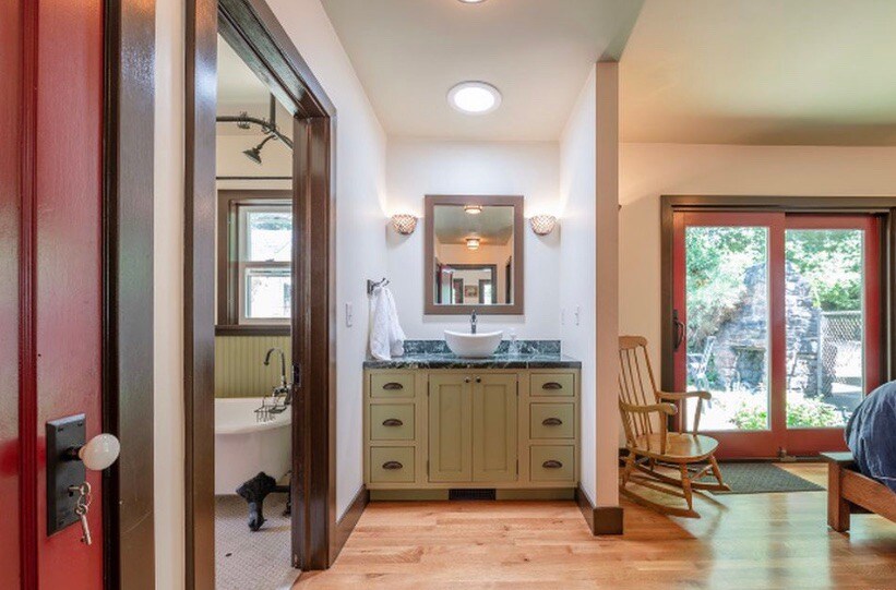 A bathroom area is visible with a combination of green cabinetry and a round mirror above a modern sink. Natural light enters through a skylight, and a rocking chair is positioned near a window, overlooking a private yard with foliage.