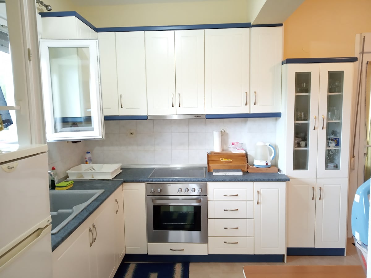 A modern kitchen is showcased, featuring white cabinets and blue accents. Stainless steel appliances, including an oven and a kettle, are visible. A sink is positioned under a window, with light-colored countertops and tiled backsplash enhancing functionality.